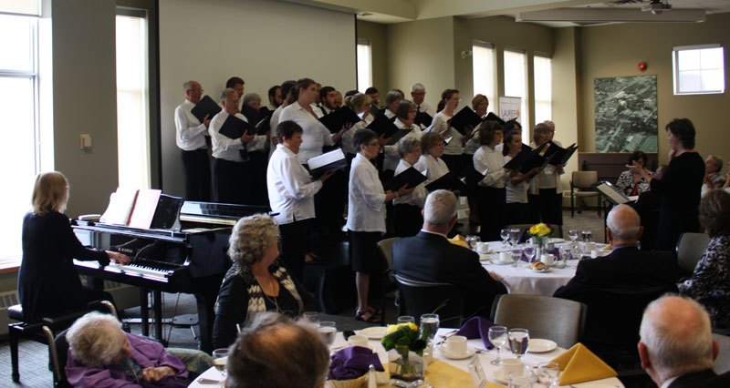 A choir singing, wearing black and white, while a person plays the piano, and others sit at tables with refreshments and watch. There are big windows letting a lot of light in.